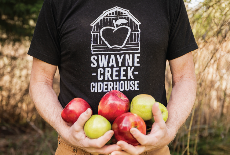 Close-up of a man's arms and chest holding a bunch of apples, wearing a Swayne Creek T-shirt