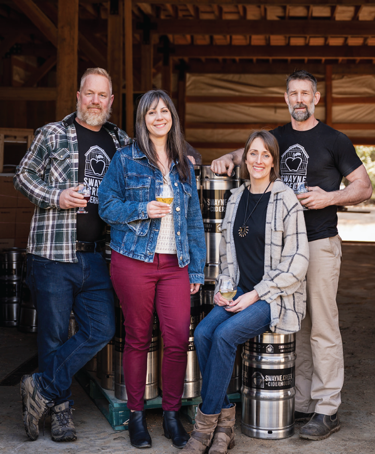 Swayne Creek Cider owners sitting on barrels, holding glasses of cider, with rustic barn elements in the background