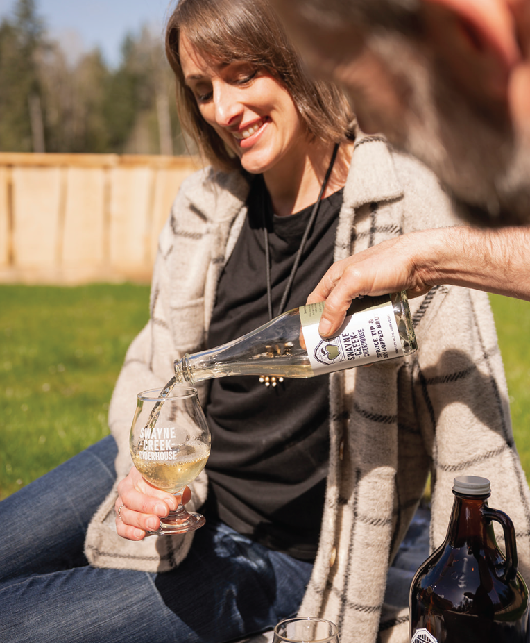 Woman sitting on a blanket outdoors while her friend pours Swayne Creek cider house into her glass.