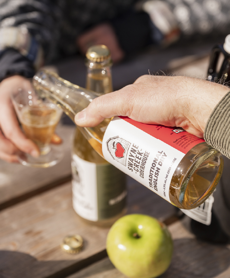 One hand pouring Swayne Creek cider into a small glass for someone else. Another bottle of cider and an apple are on the table.